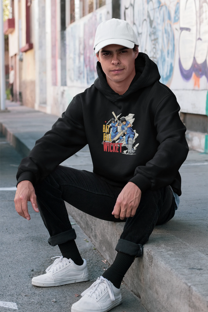 Young man seated on an urban curb, wearing a black hoodie featuring a 'Bat Ball Wicket' graphic with animated cricket players, paired with black jeans and white sneakers. He also wears a white baseball cap, against a backdrop of vibrant graffiti.