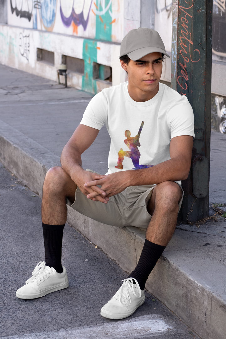 Young man in a casual setting, squatting by a graffiti-covered wall. He's wearing a light-colored T-shirt with a vibrant cricket player print, khaki shorts, white sneakers, black socks, and a gray baseball cap.